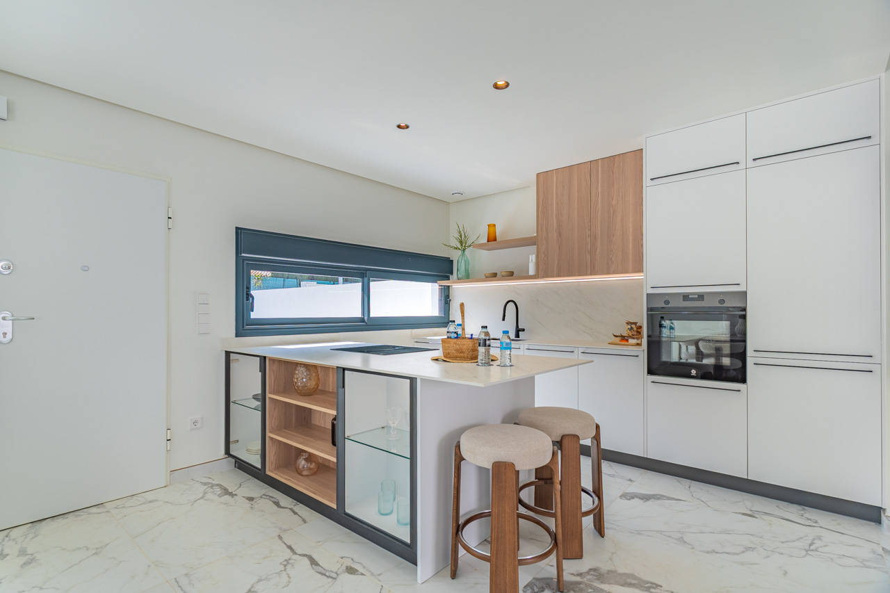 Modern open-concept kitchen featuring a central island with seating, white cabinetry, and wood accents. It includes integrated appliances and marble-effect porcelain flooring.
