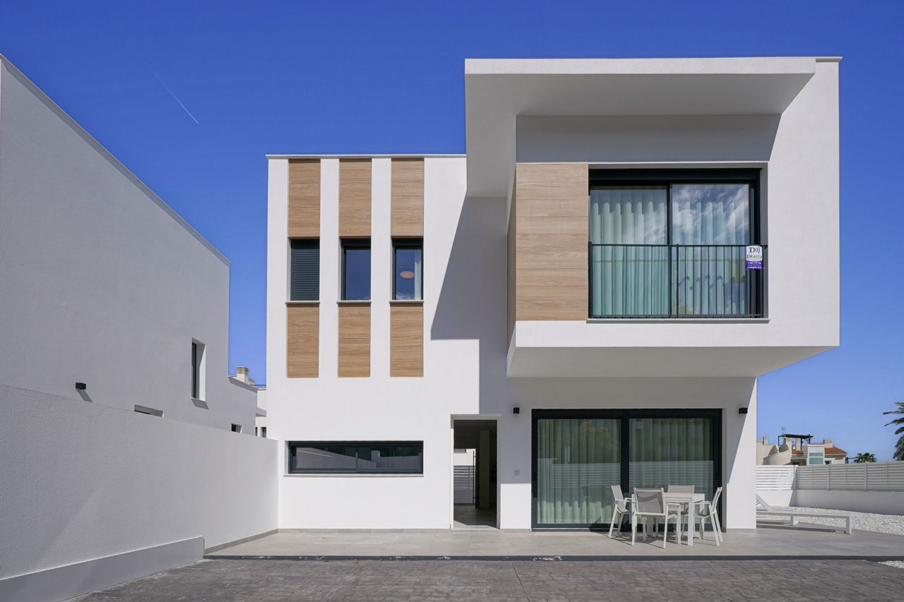 Modern villa exterior featuring a white facade with wood-effect accents, a spacious ground-floor terrace with dining furniture, and a cantilevered upper level.