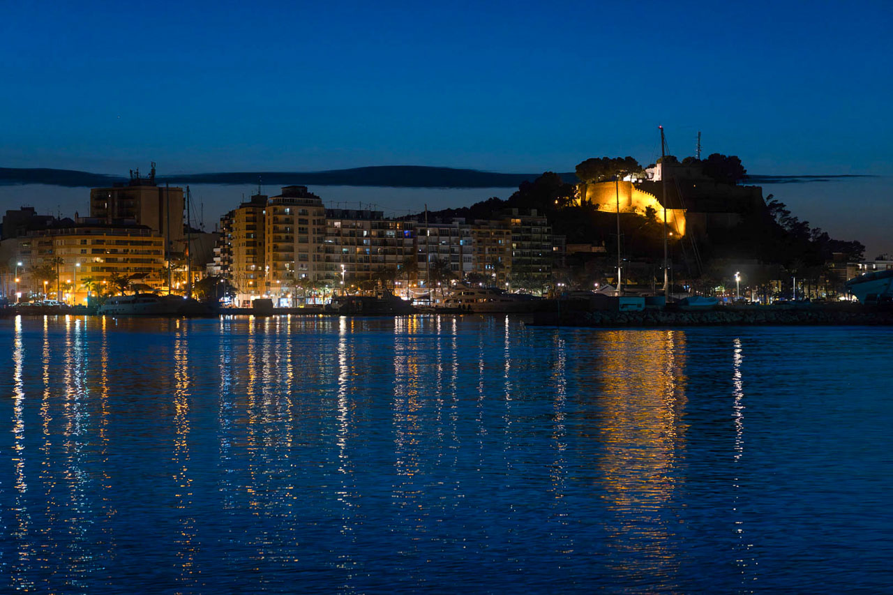 Night view of Dénia harbor featuring the illuminated castle on the hill and city lights reflecting across the calm Mediterranean waters.
