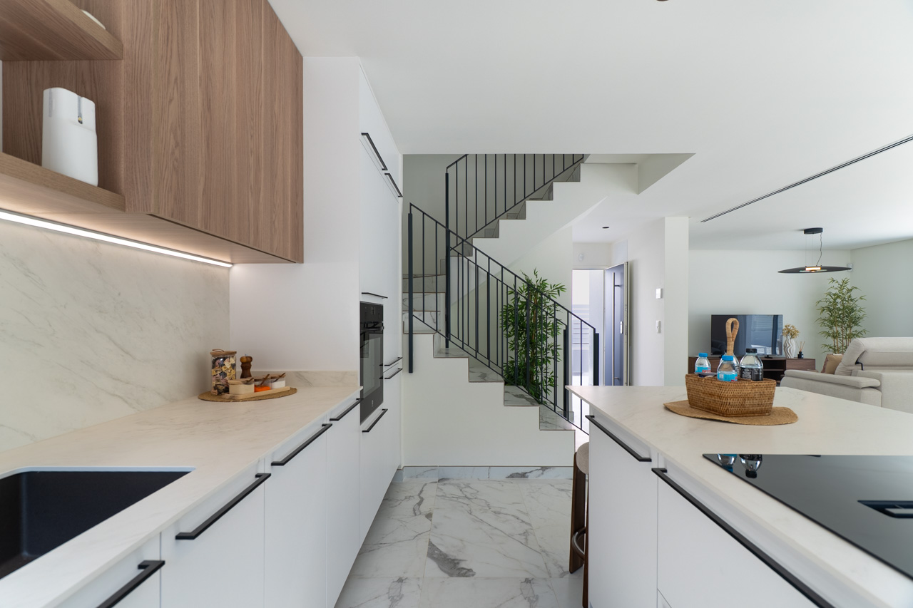 Open-concept kitchen featuring white cabinetry, marble-effect countertops, and wooden upper units with integrated LED lighting. A minimalist staircase leads to the upper floor.