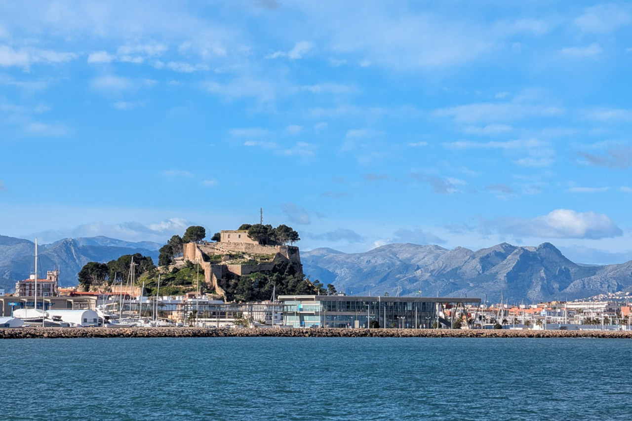 Panoramic view of Dénia harbor showcasing the historic castle and the Montgó mountain range under a clear blue sky from the Mediterranean Sea.
