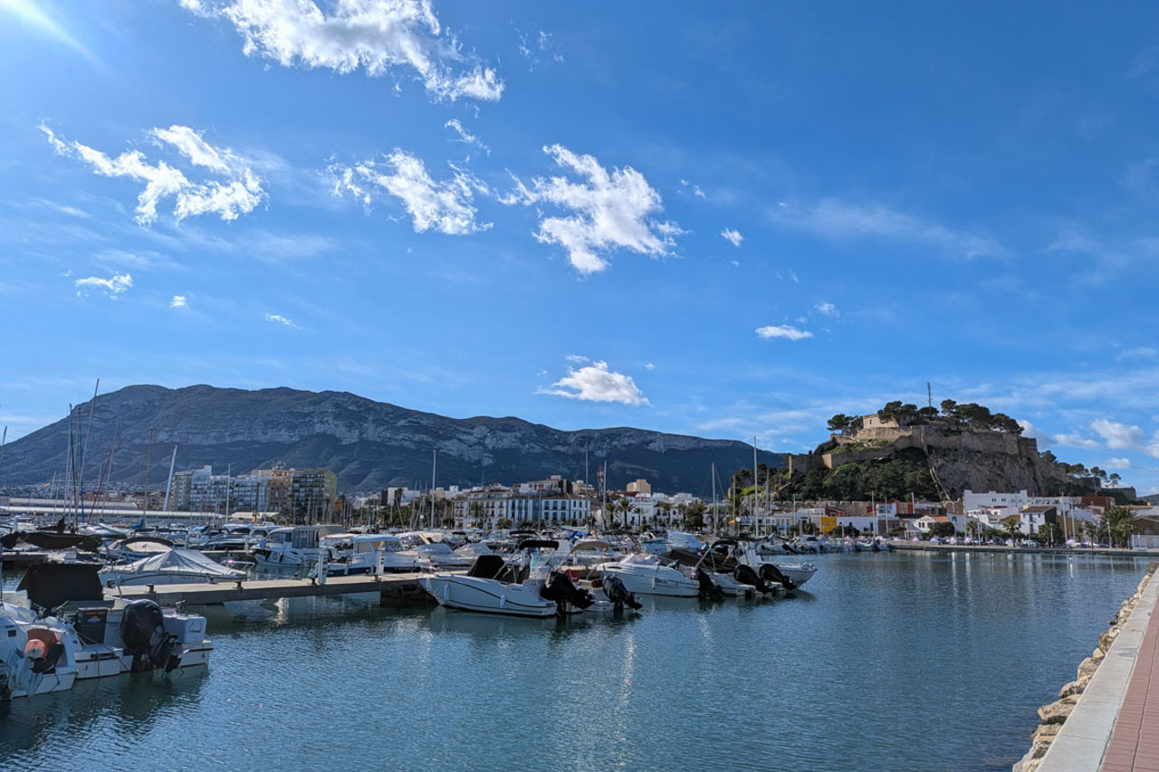 Panoramic view of Dénia's marina featuring docked boats, the historic hilltop castle, and the Montgó mountain range in the background.