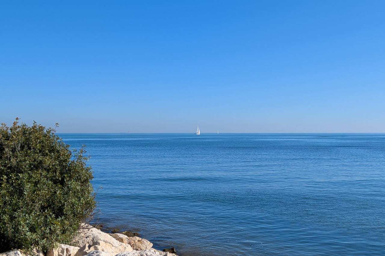 Panoramic view of the Mediterranean Sea from Dénia's rocky coastline, featuring sailboats on the horizon under a clear blue sky.