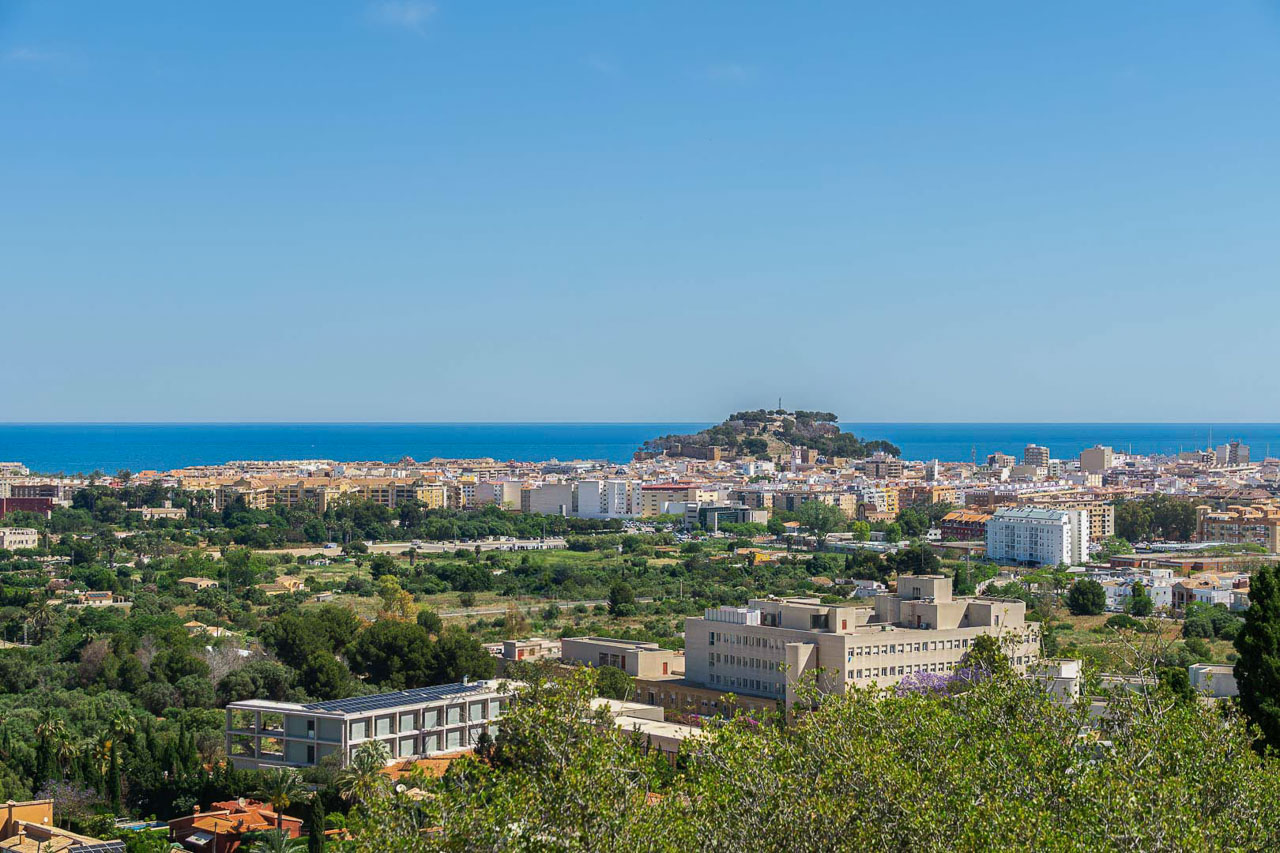 Panoramic views of Dénia featuring the historic castle and the Mediterranean Sea on the horizon. The elevated perspective showcases the urban landscape and coastal proximity.