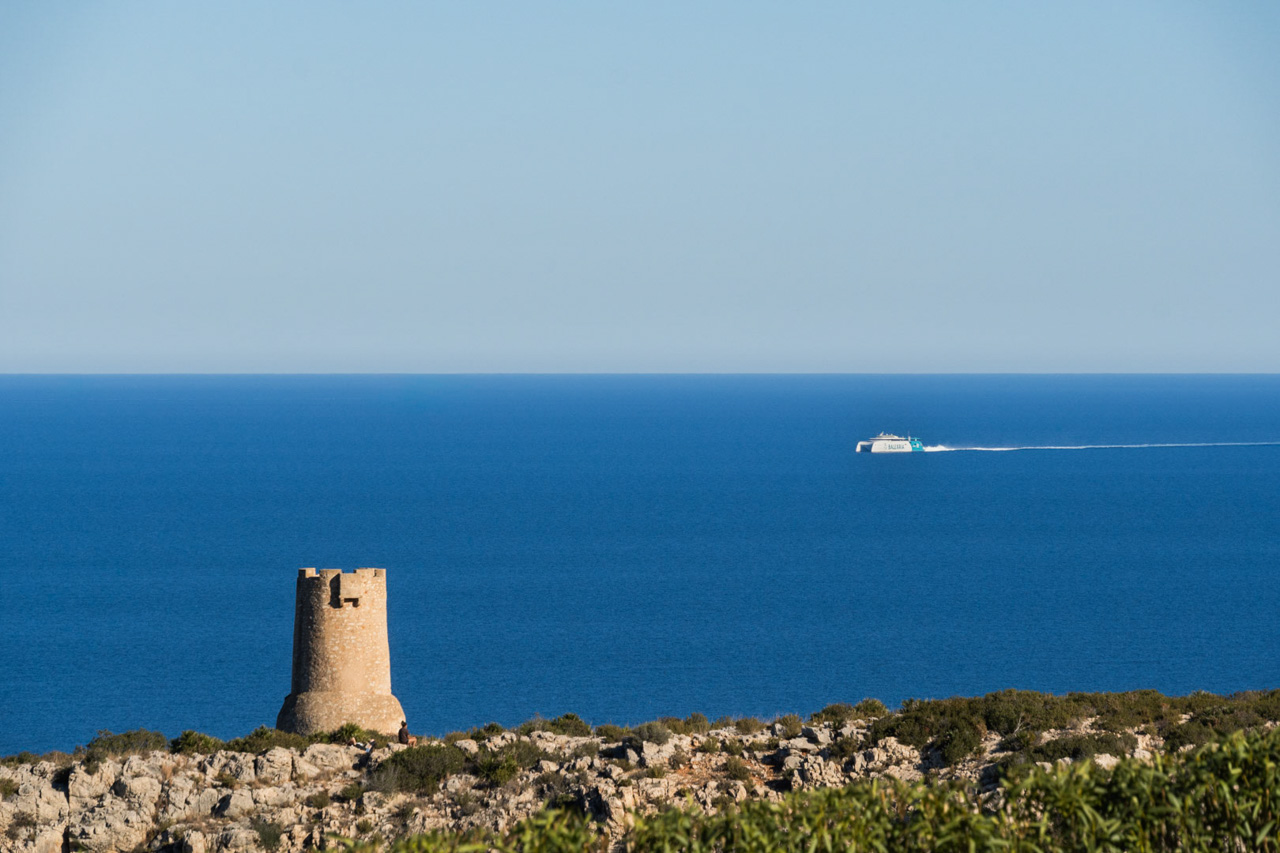 Panoramic views of the Las Rotas coastline in Dénia, featuring the historic Torre del Gerro watchtower and a Balearia ferry crossing the Mediterranean Sea.