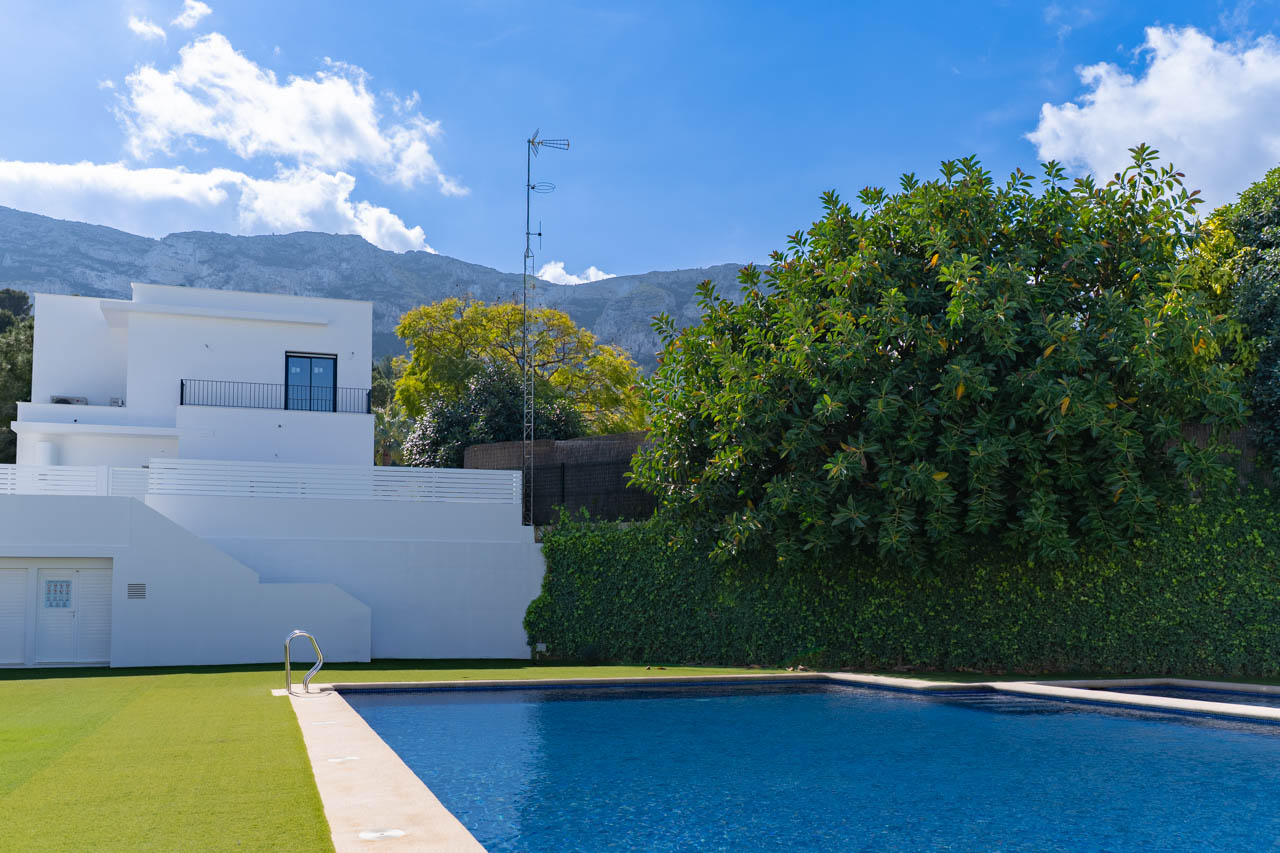 Rectangular swimming pool with stone coping and artificial lawn, set against a minimalist white facade with views of the Montgó mountain range.