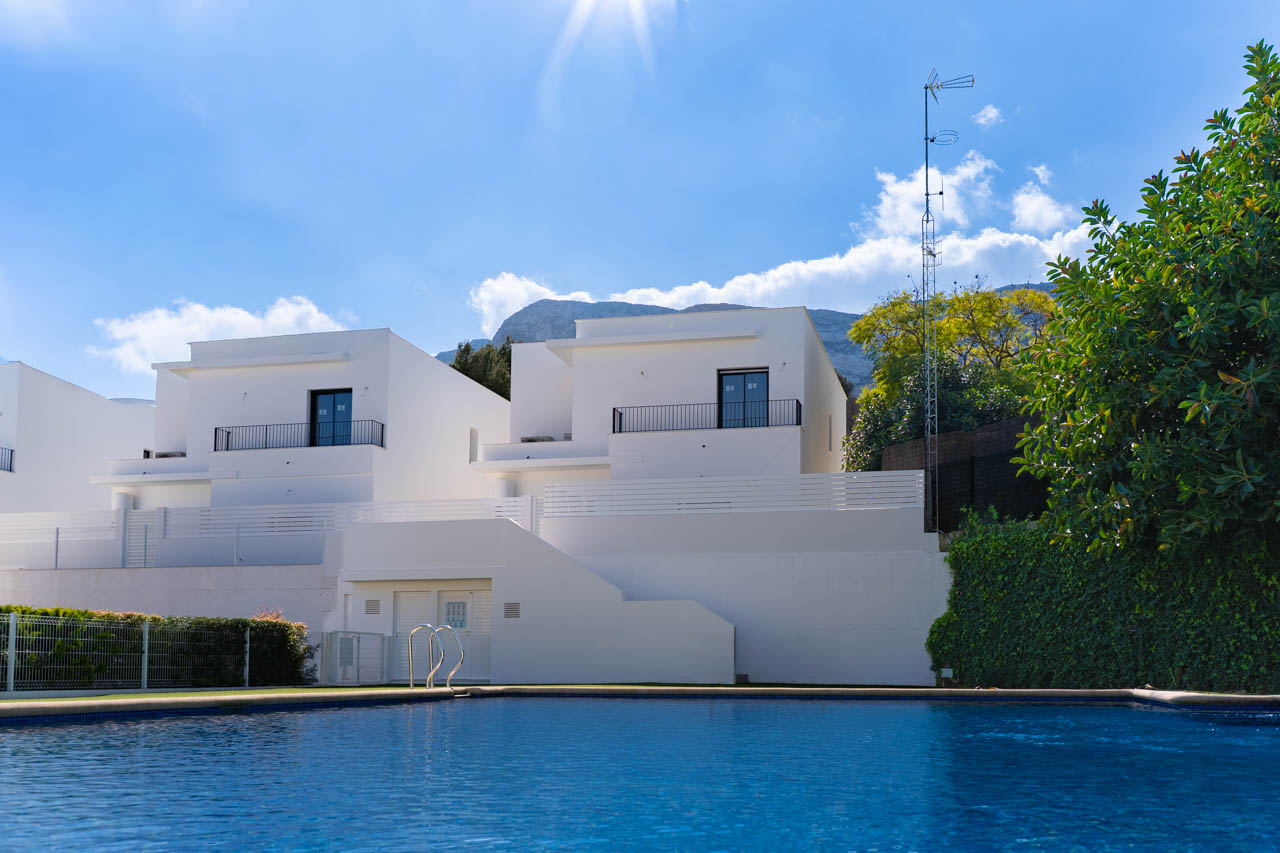 View of the modern villas from the communal pool, showcasing white minimalist facades and the Montgó mountain range under a clear blue sky.
