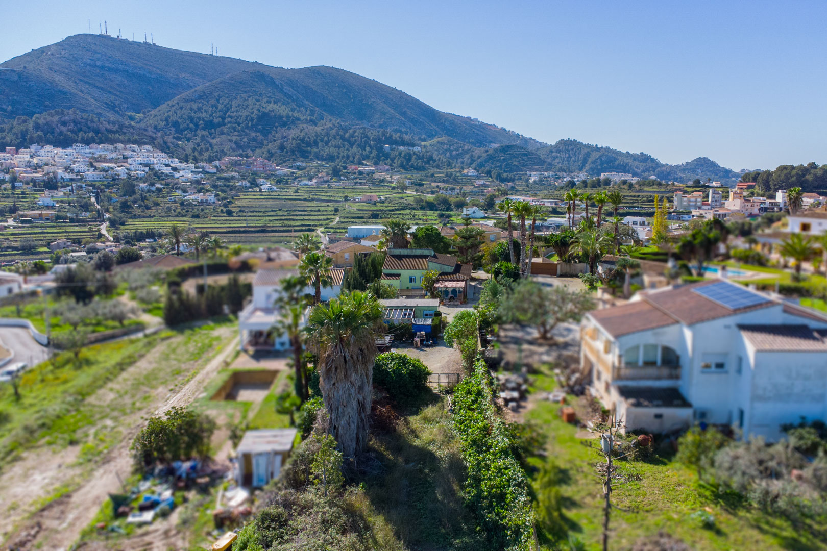 Aerial view of the rustic plot showing the green-facade main house, prefab unit, and surrounding terraced valleys with mountain views.