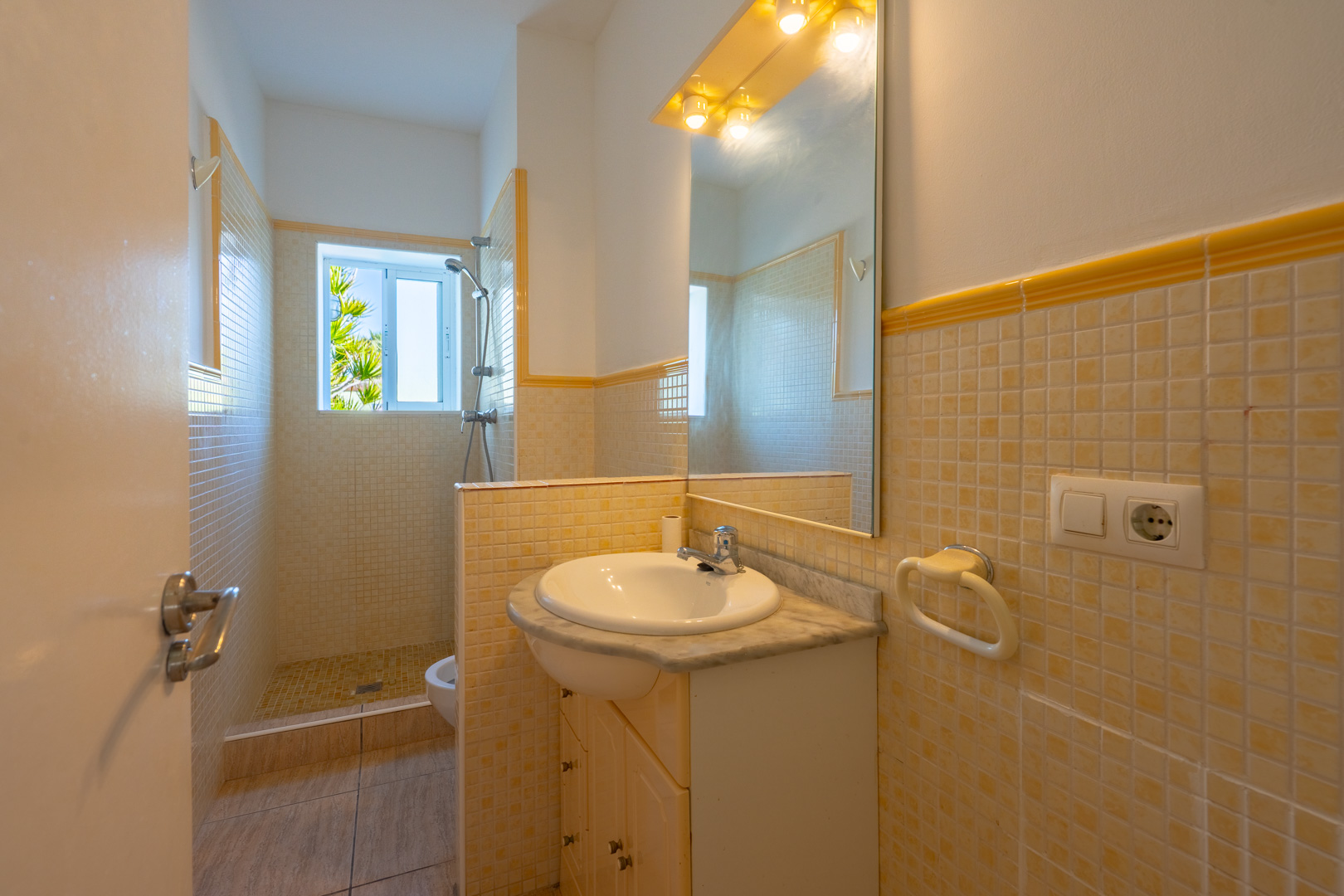 Bathroom featuring a walk-in mosaic shower, marble-top vanity, and a large mirror with warm lighting. A window provides natural light and garden views.