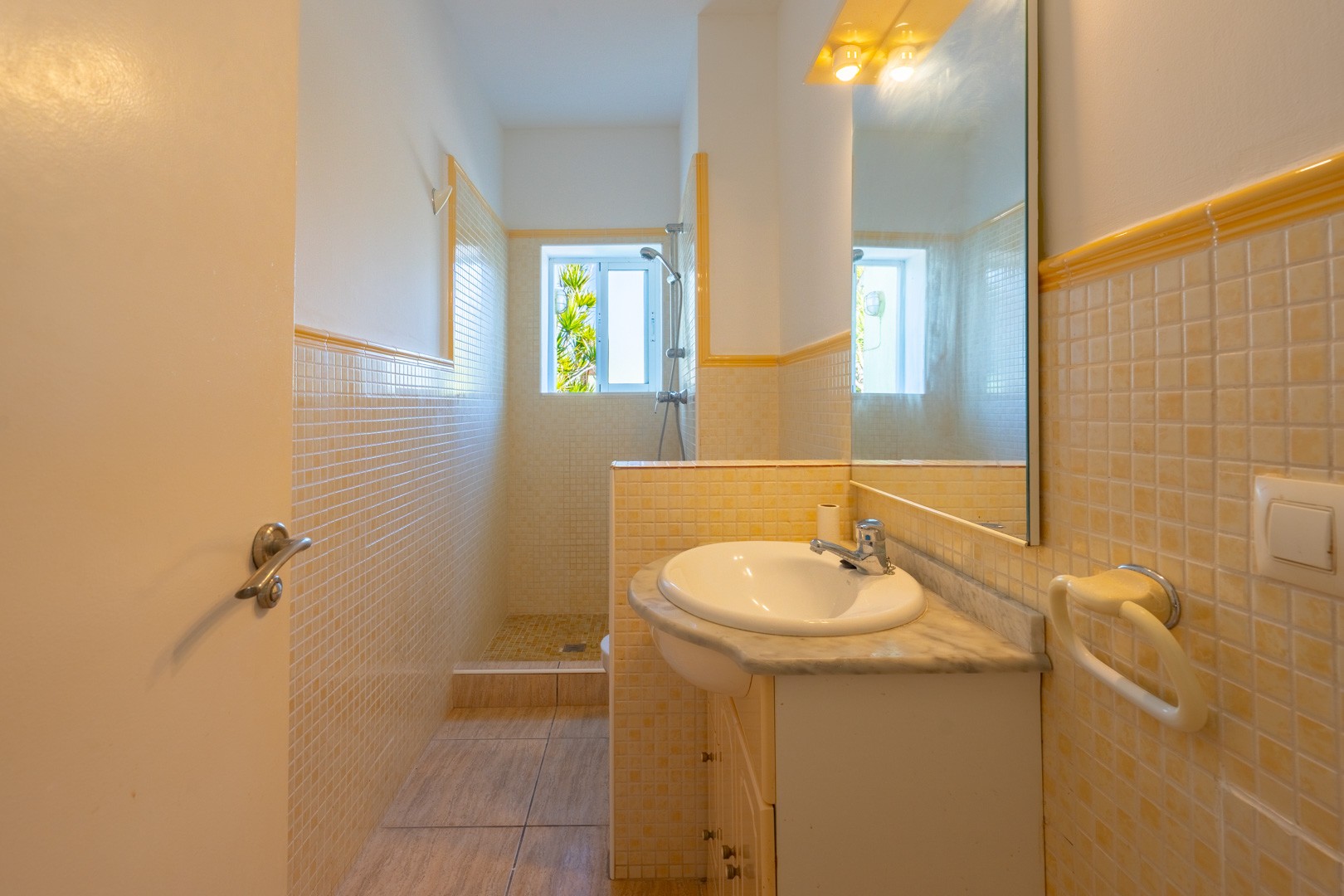 Bathroom featuring a walk-in shower, cream and yellow mosaic tiling, a marble-topped vanity unit, and a window providing natural light.