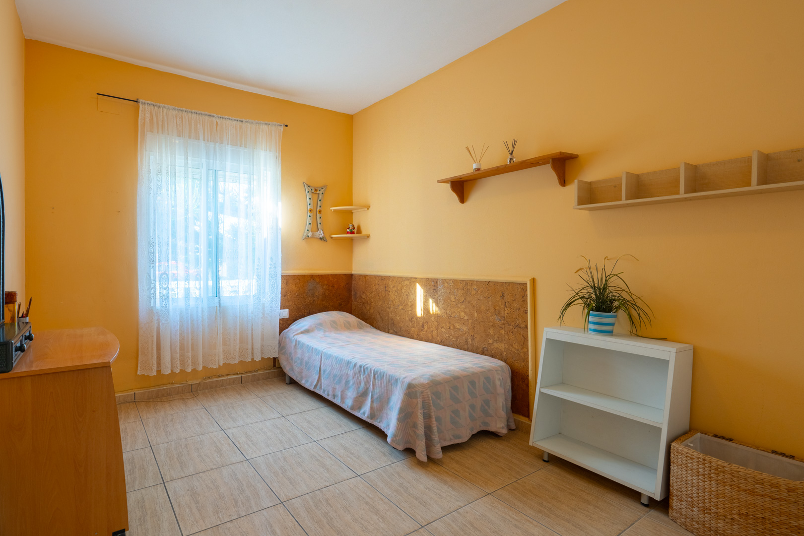Bedroom featuring ochre walls, ceramic tile flooring, a single bed, and wooden shelving. A window with lace curtains provides soft natural light.