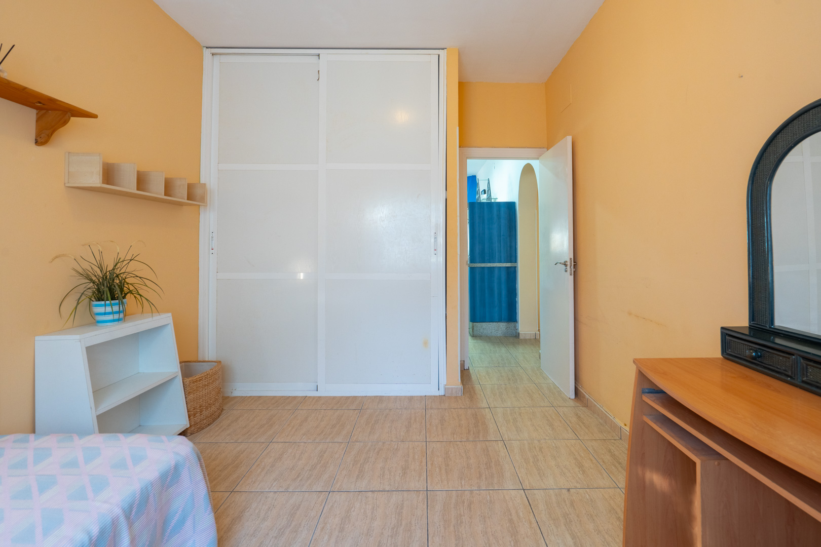 Bedroom featuring peach-colored walls, a large white built-in sliding wardrobe, ceramic tile flooring, and a wooden vanity with an arched mirror.