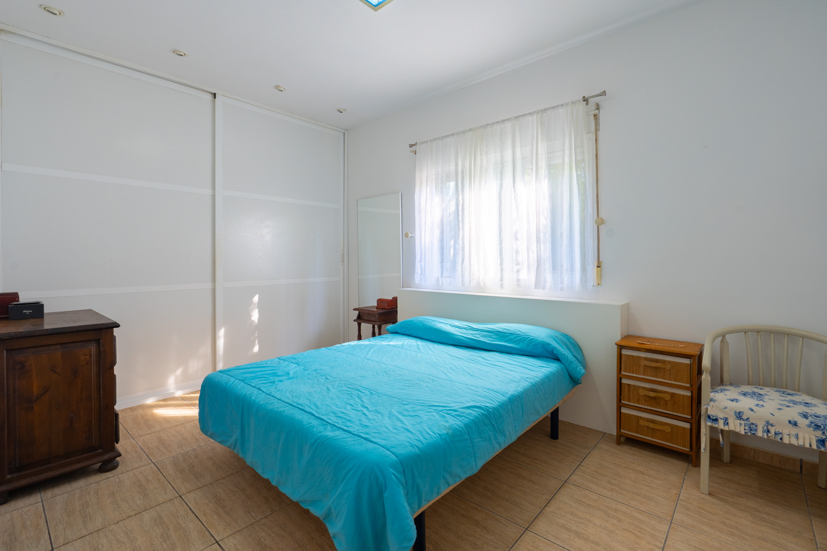 Bright bedroom featuring white built-in sliding wardrobes, tiled flooring, natural light through sheer curtains, and classic wooden furniture.