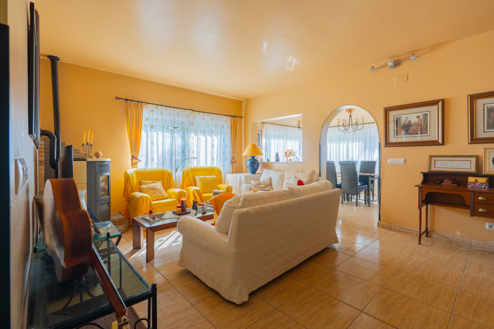 Bright living room featuring ochre walls, a wood-burning stove, and tiled flooring. An arched doorway leads to the dining area, complemented by natural light.