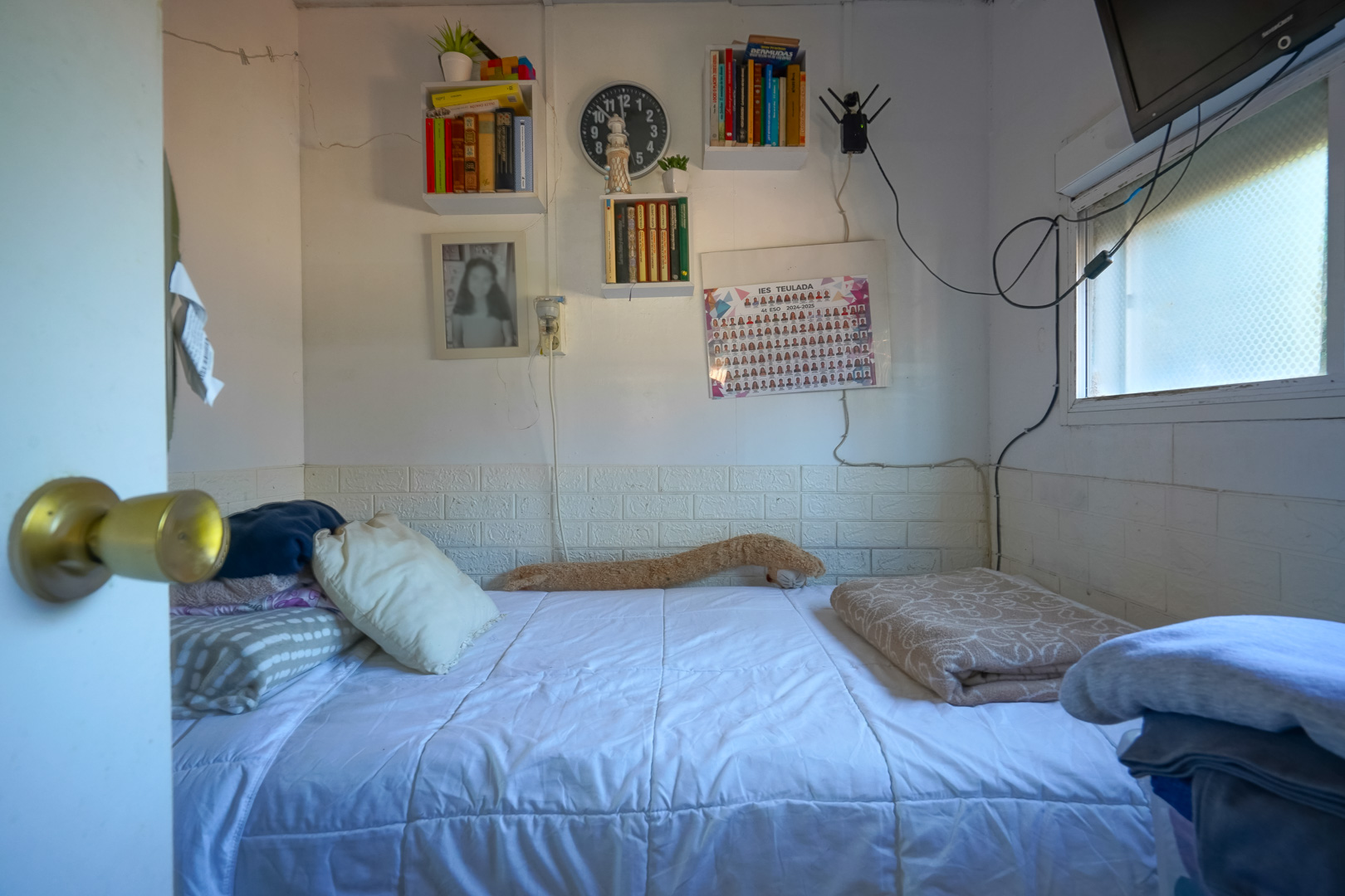 Compact bedroom featuring a single bed, wall-mounted bookshelves, and a side window. The walls include white brick-effect wainscoting and functional storage.