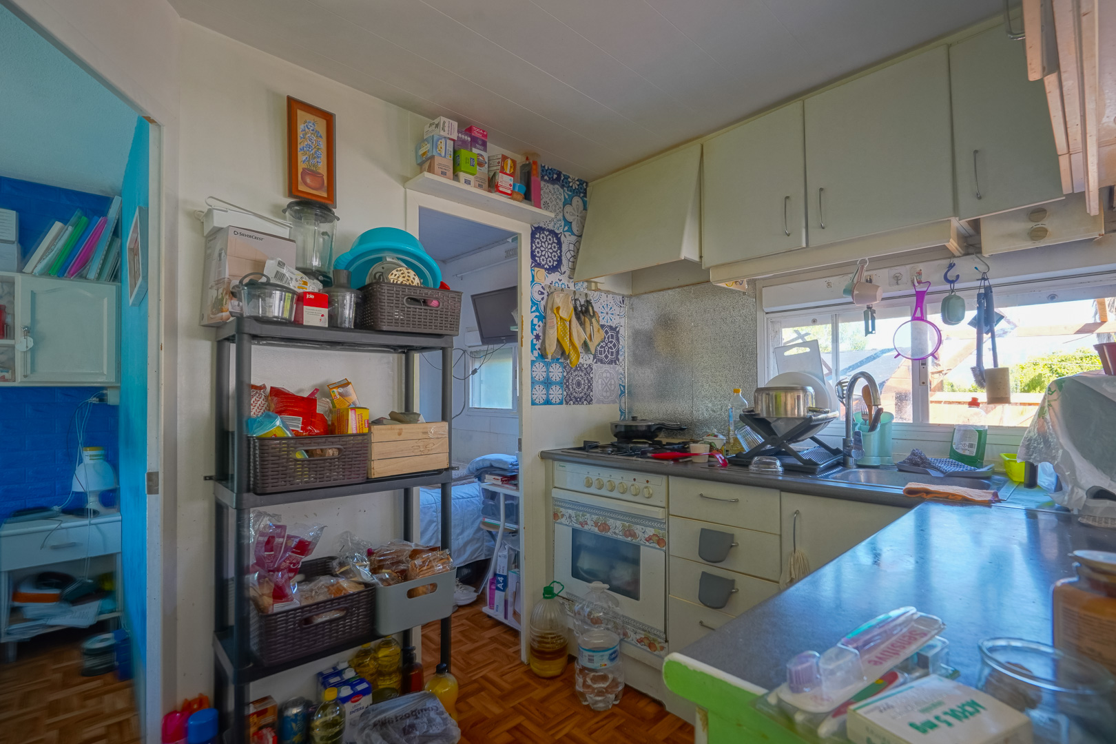 Compact kitchen in the guest house featuring white cabinetry, decorative mosaic tiles behind the stove, and practical storage shelving under natural light.
