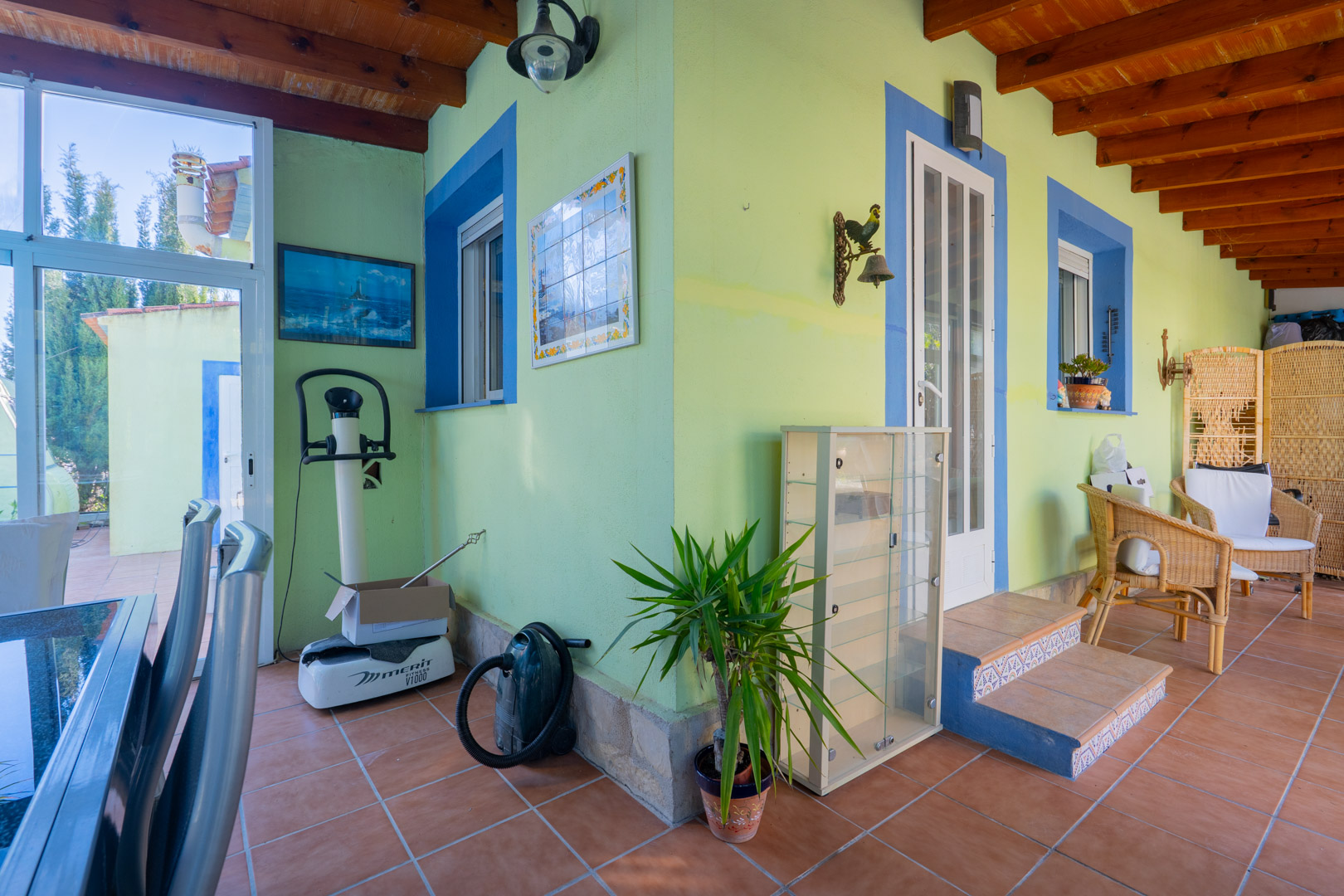 Covered terrace featuring exposed wooden beams, vibrant green walls with blue accents, terracotta flooring, and traditional wicker seating.