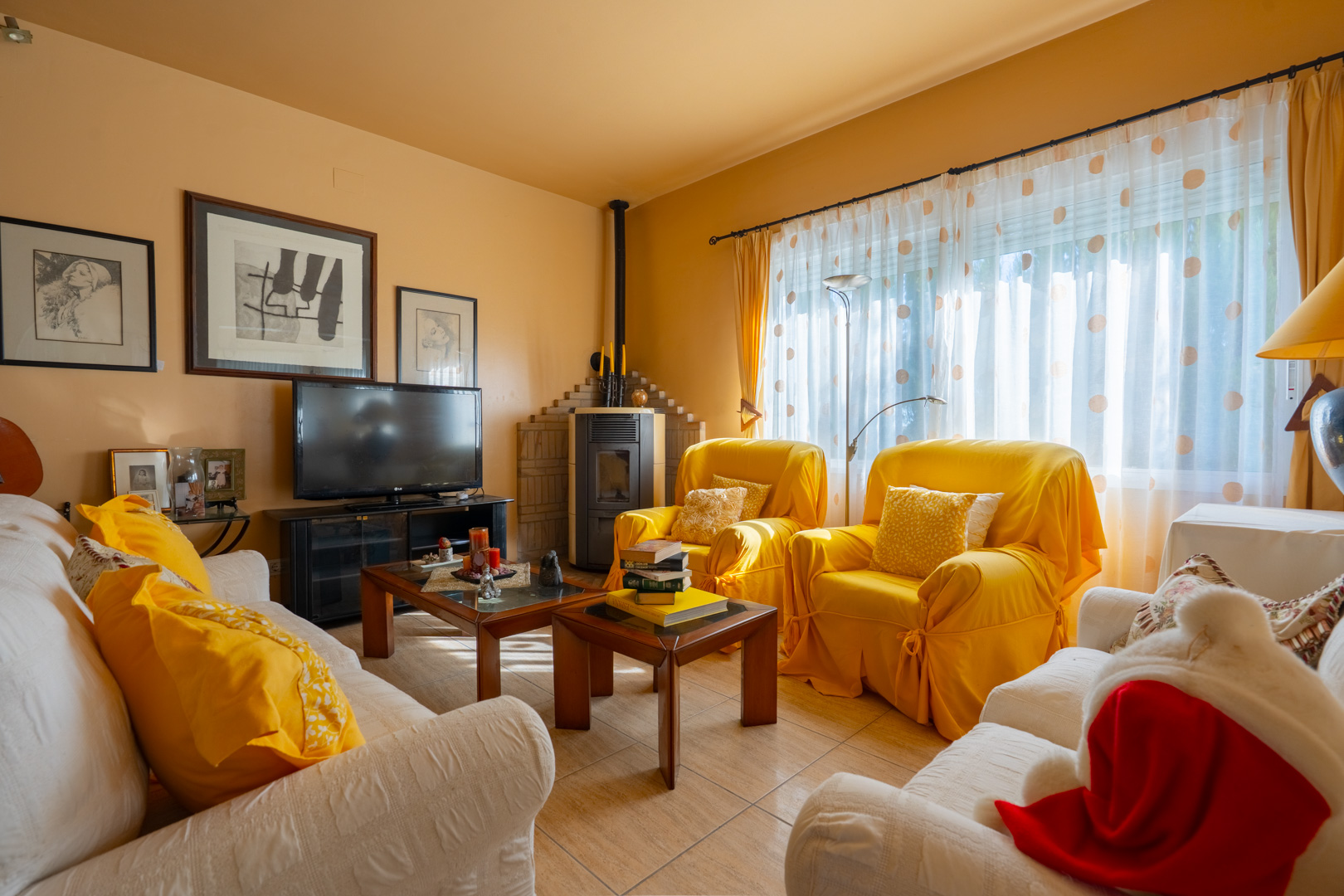 Cozy living room featuring a corner wood-burning stove, yellow and white upholstered seating, and natural light through patterned curtains.