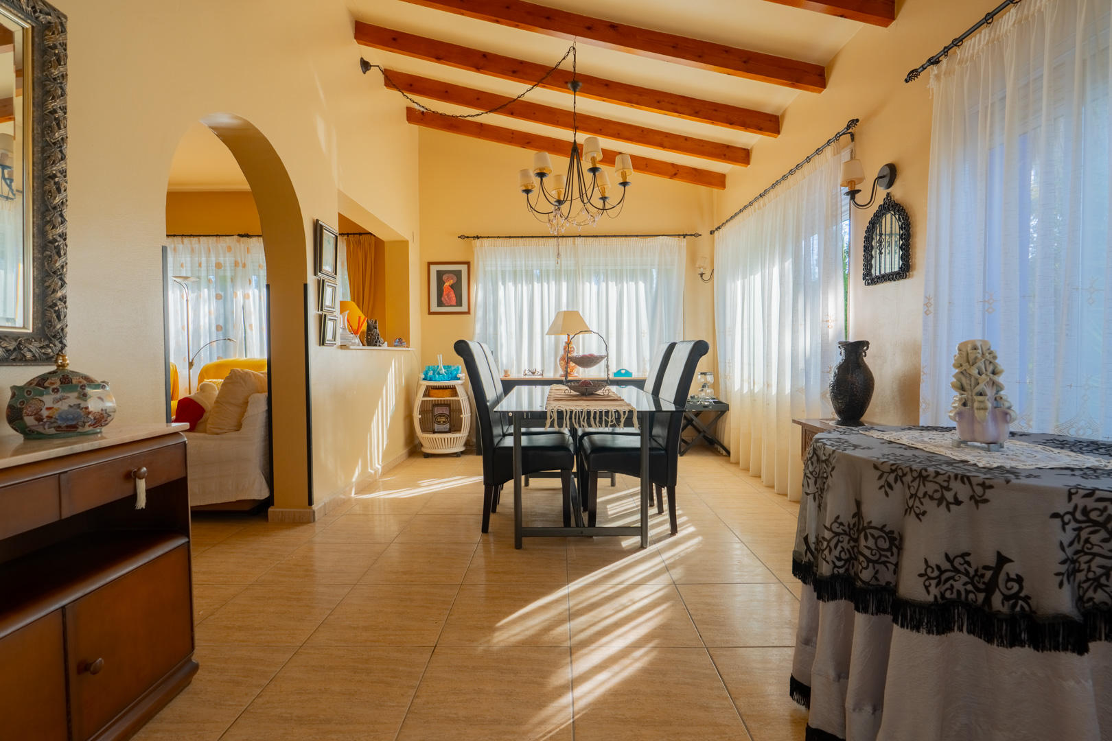 Dining area featuring high ceilings with exposed wooden beams, tiled flooring, and an elegant archway connecting to the adjacent living space.