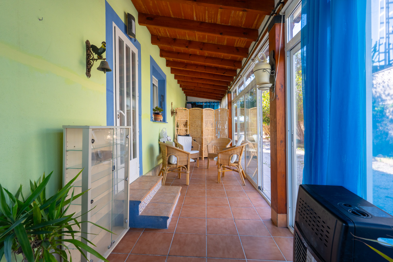 Enclosed naya featuring exposed wooden beams, terracotta tile flooring, and wicker seating, integrated with the outdoors via large glass sliding doors.