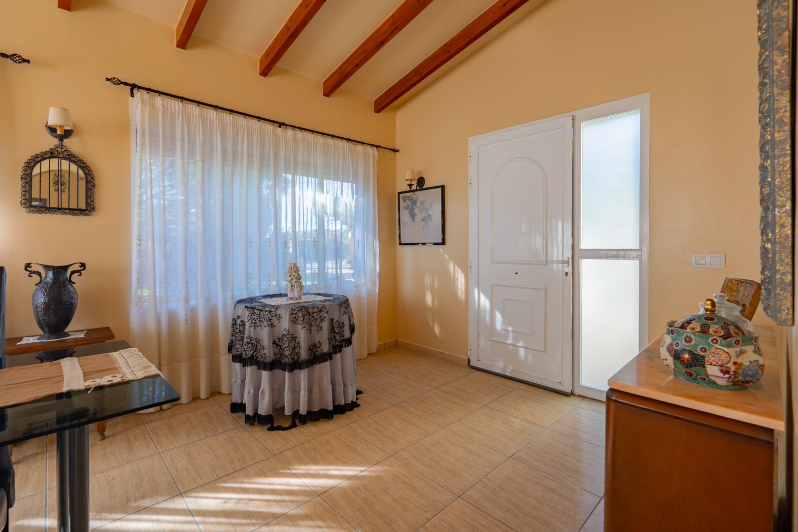 Entrance hall featuring high ceilings with exposed wooden beams, ceramic tile flooring, and a white front door with a frosted glass side panel.