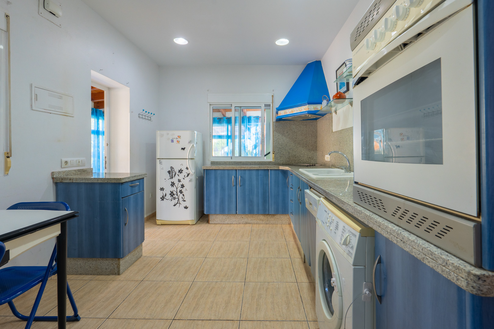Equipped kitchen featuring blue cabinetry, grey granite countertops, and integrated appliances. Includes recessed lighting, ceramic flooring, and natural light from the window.