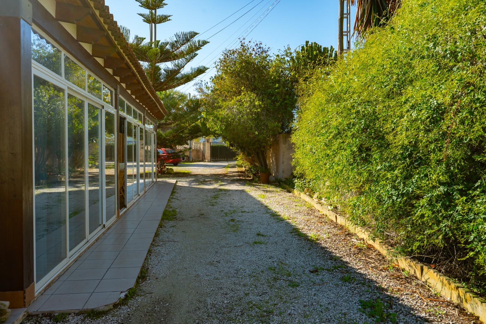Exterior view of the glazed naya with wooden beams, adjacent to a gravel driveway bordered by mature Mediterranean greenery.