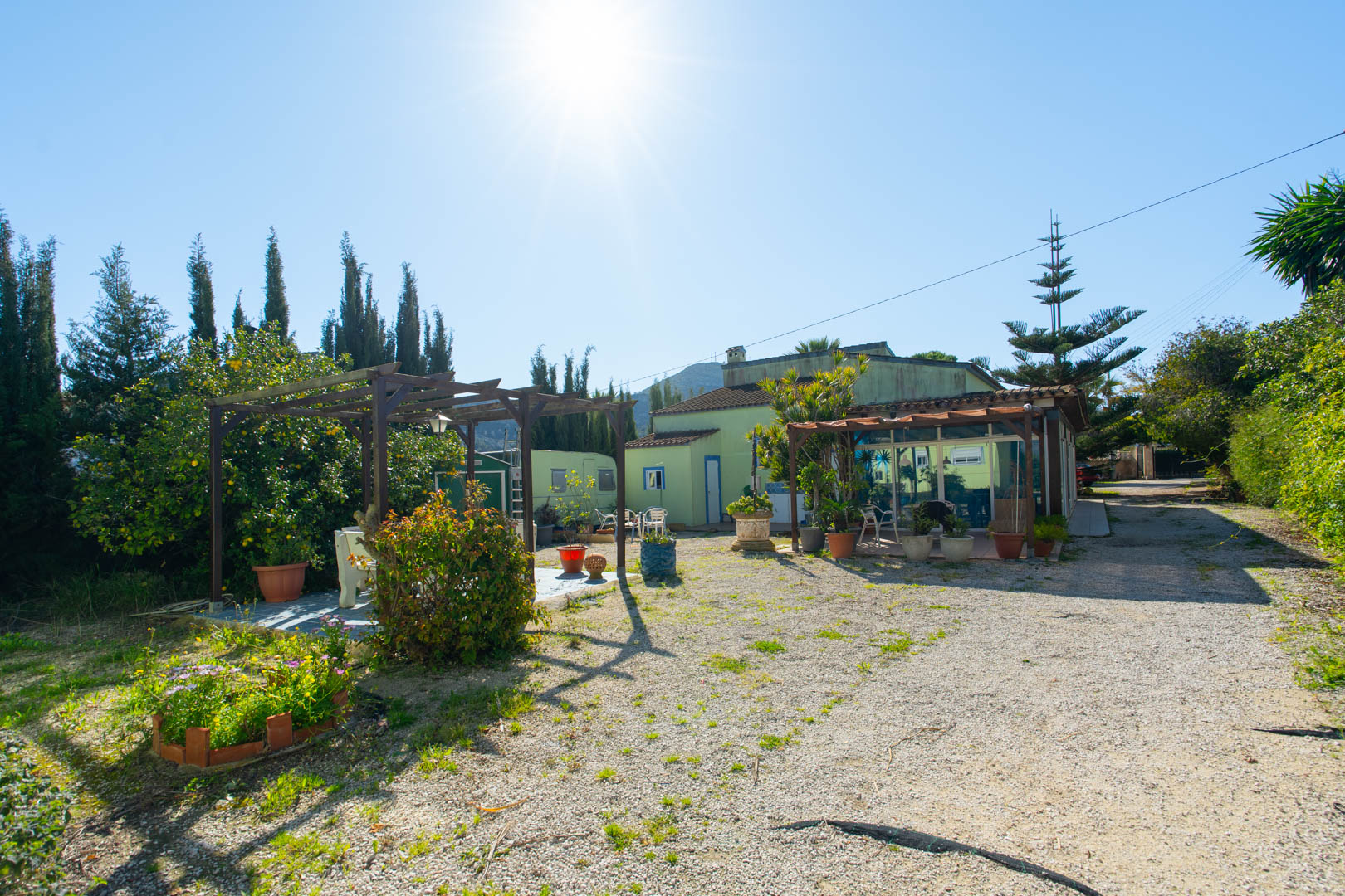 Exterior view of the rustic garden featuring a wooden pergola, seating area, and the main house with a glass-enclosed naya and gravel driveway.