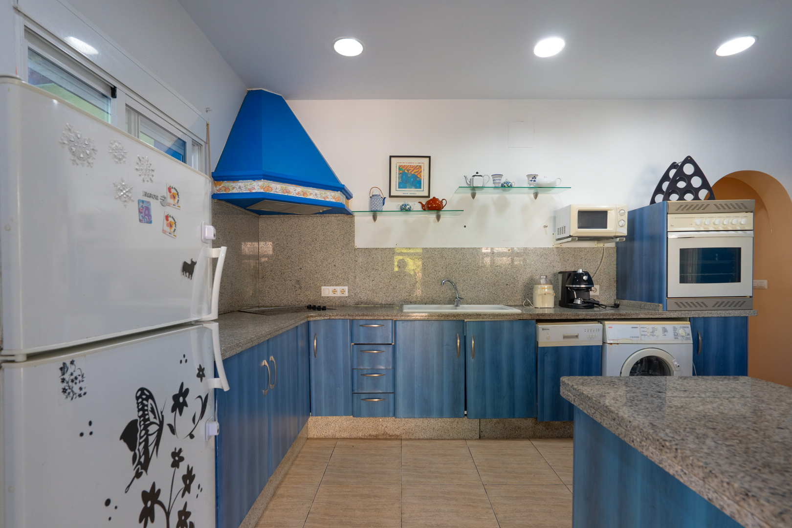 Functional kitchen featuring blue cabinetry, grey granite countertops, and a matching backsplash. Equipped with a blue extractor hood, integrated appliances, and recessed lighting.