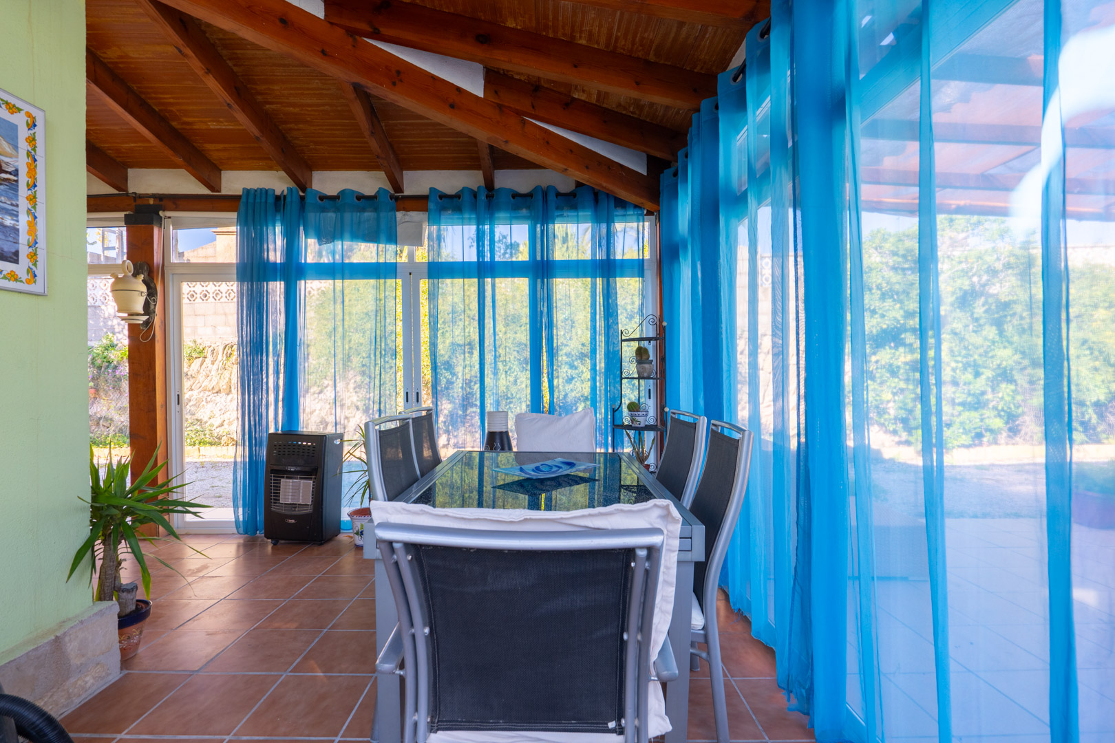 Glazed naya dining area featuring exposed wooden ceiling beams, terracotta flooring, and blue sheer curtains overlooking the garden.