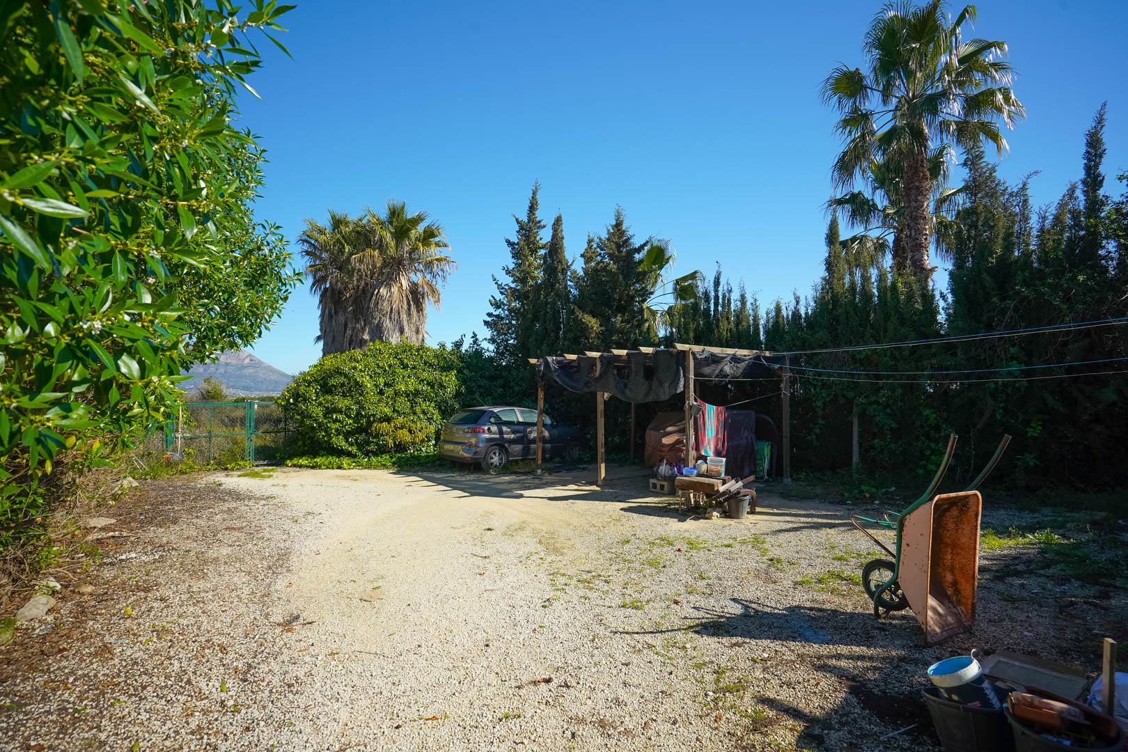 Gravel parking area featuring a rustic wooden carport, surrounded by mature palm trees and cypresses with distant views of the Montgó mountain.