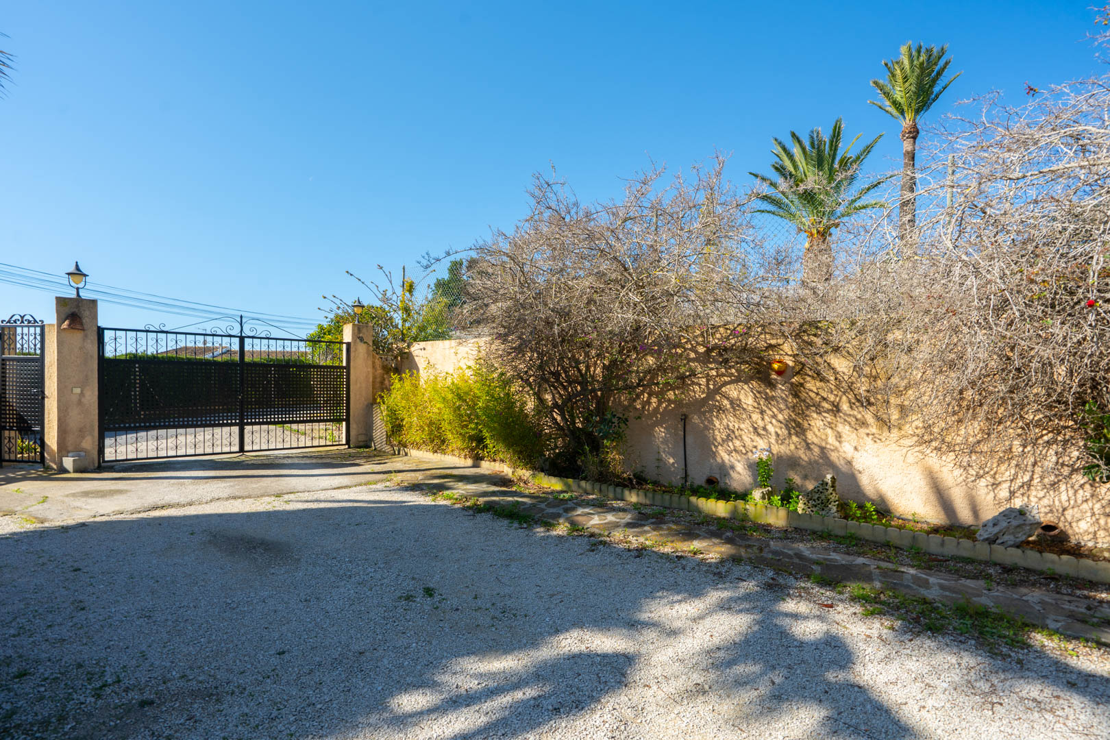Main entrance featuring a black wrought-iron gate, a gravel driveway, and a perimeter wall lined with Mediterranean plants and palm trees.