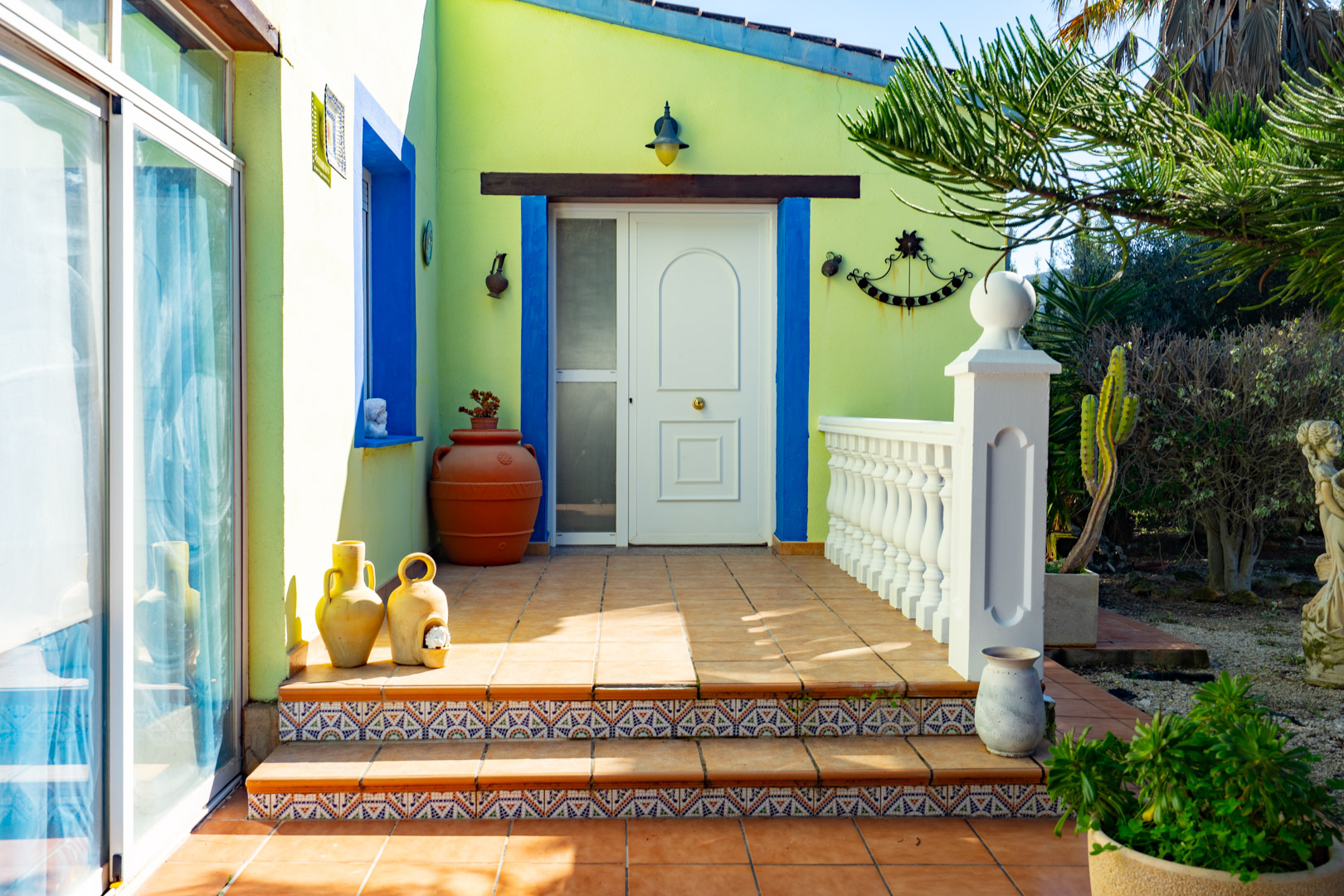 Main entrance with a white door, blue window frames, and decorative tiled steps. The terracotta terrace features a white balustrade and traditional Mediterranean pottery.