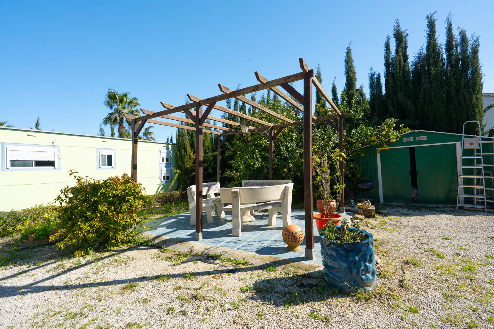 Outdoor seating area with a wooden pergola and stone benches on a tiled base, located between the green storage shed and the prefabricated guest house.