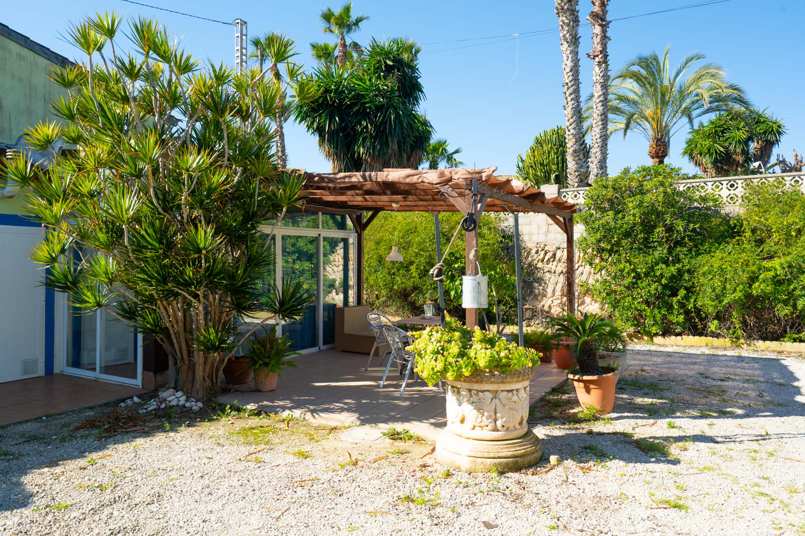 Outdoor terrace featuring a wooden pergola, dining area, and lush Mediterranean greenery including palms and succulents in decorative stone planters.