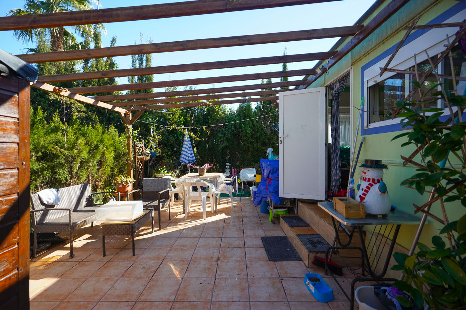 Outdoor terrace of the prefabricated unit featuring ceramic tiling, a wooden beam pergola, and a seating area enclosed by mature hedges.