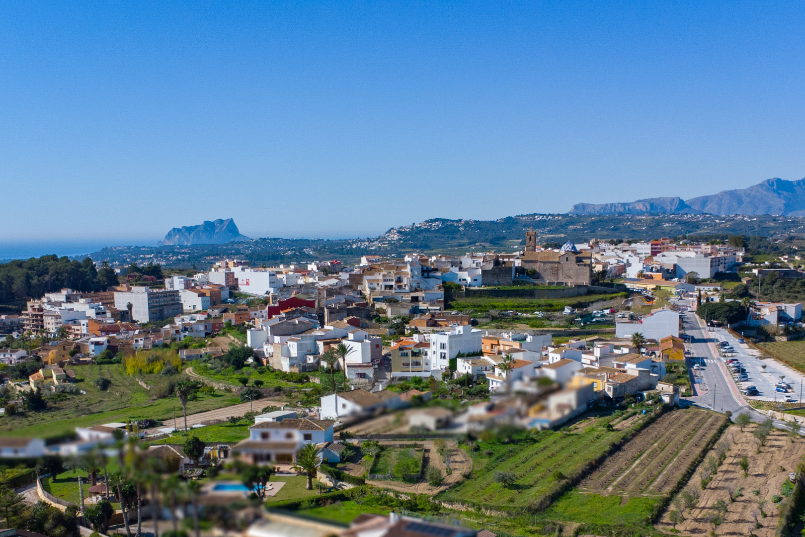 Panoramic aerial view of Benitachell featuring the historic town center, the church, and the Peñón de Ifach against the Mediterranean coastline.