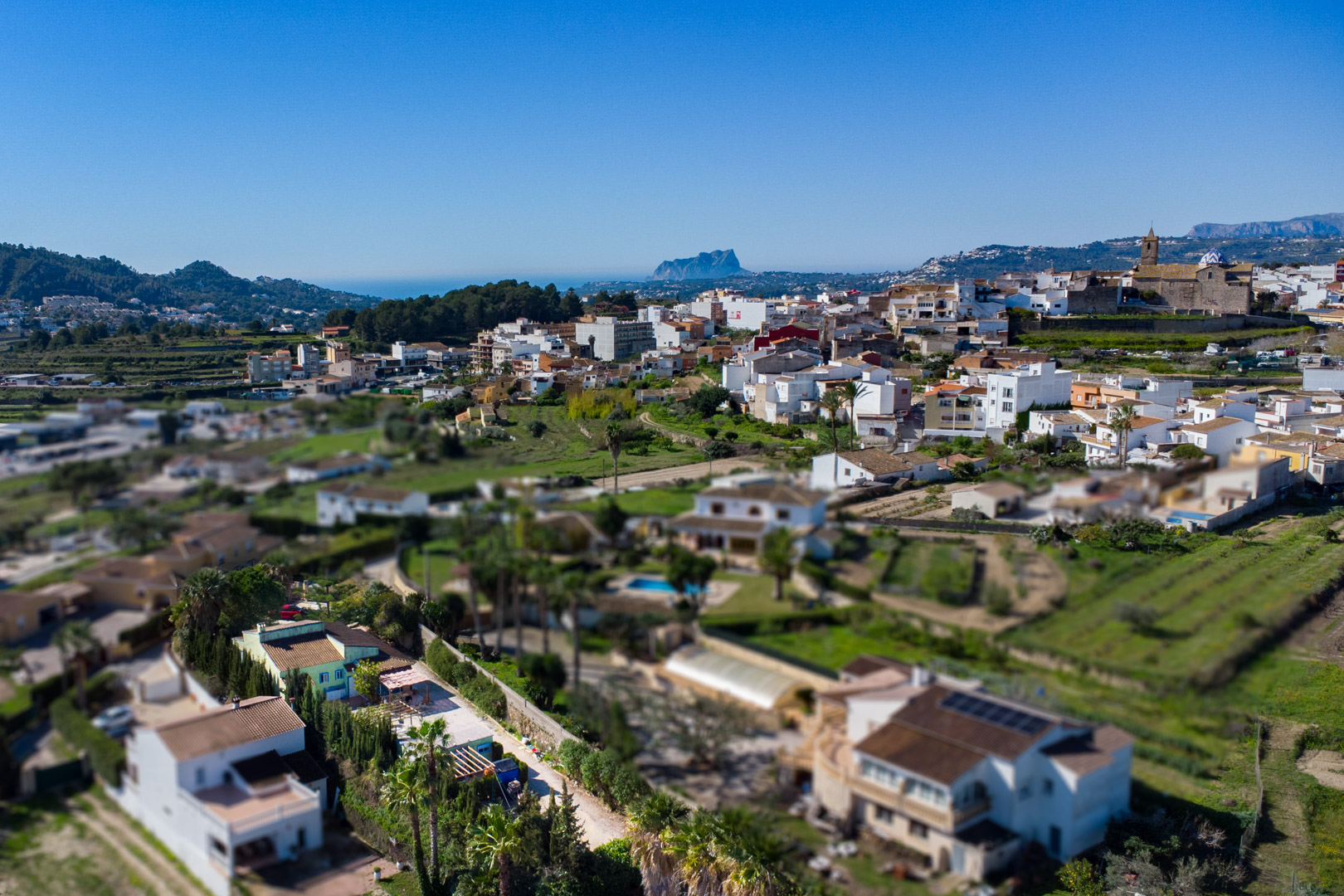 Panoramic aerial view of Benitachell featuring the Peñón de Ifach on the horizon. The image shows the town center, the parish church, and the surrounding Mediterranean valley.