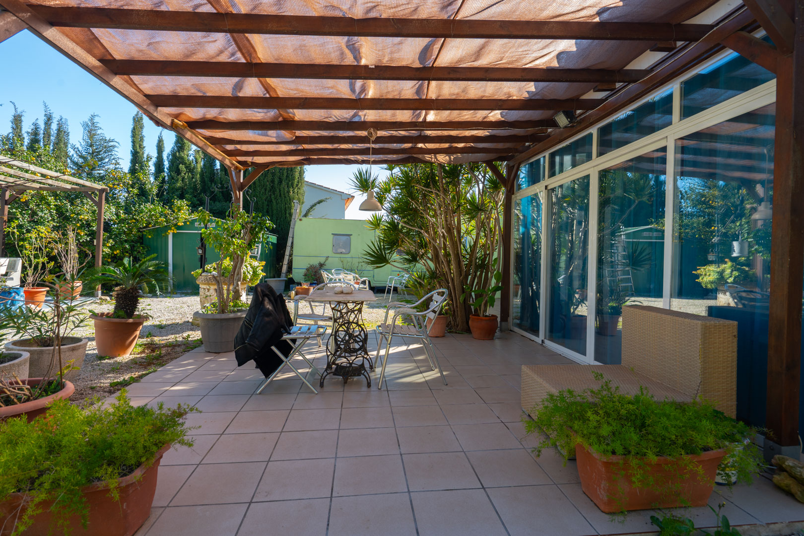 Paved terrace featuring a wooden pergola with mesh shading, outdoor seating, and various potted plants overlooking the Mediterranean garden area.