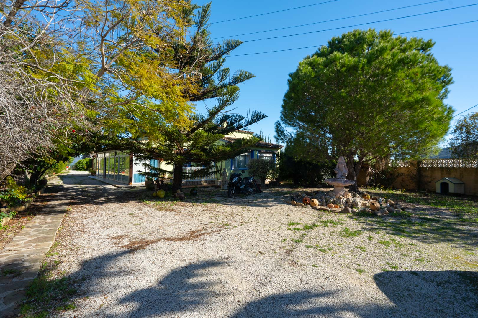 Spacious gravel entrance area featuring mature pine trees and a stone fountain, leading to the main villa with its glass-enclosed naya.