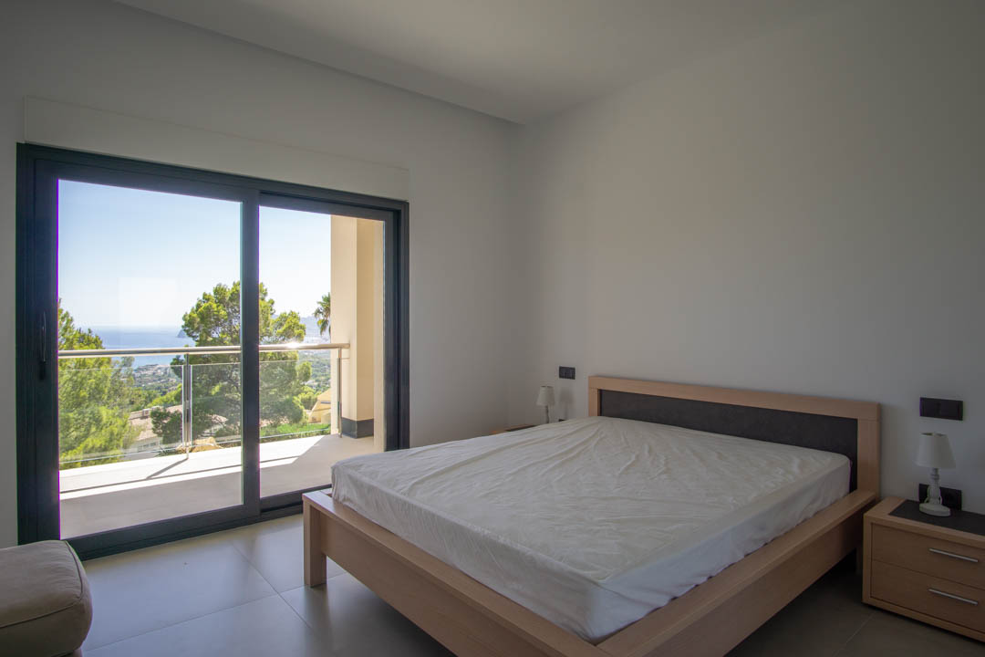 Bright bedroom featuring a light wood bed frame, direct access to a private balcony, and clear views of the Mediterranean coastline and surrounding pine trees.