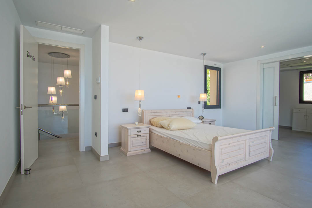 Bright bedroom featuring a light wood bed frame, matching nightstands, and pendant lighting. Large-format tile flooring leads to a hallway with a glass railing.