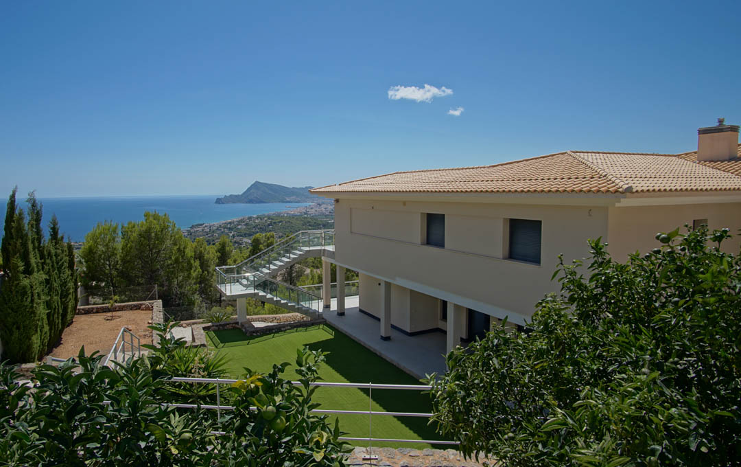 Exterior view of the villa featuring an artificial lawn, citrus trees, and glass staircase, overlooking the Altea bay and Mediterranean coastline.