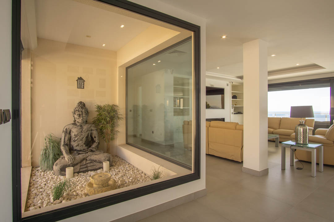 Glass-enclosed interior Zen garden featuring a Buddha statue and white pebbles, adjacent to a spacious living area with recessed lighting and sea views.