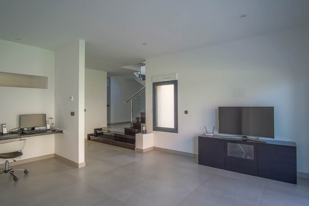Ground floor lounge featuring a built-in workspace, minimalist TV console, and a dark-tread staircase with glass balustrades and recessed lighting.