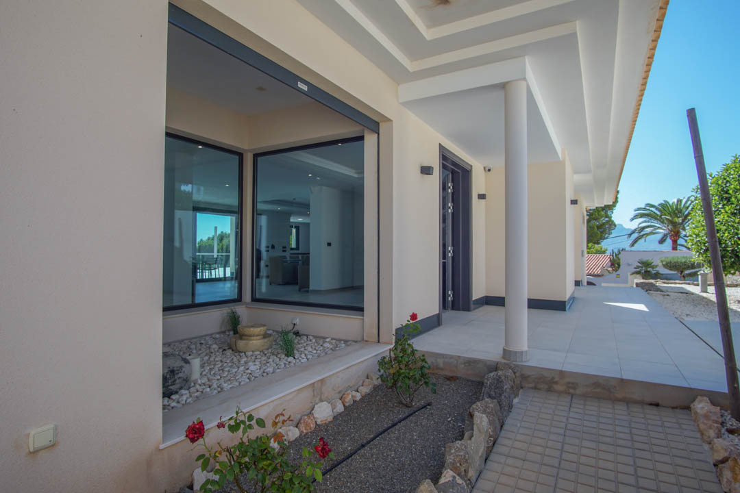 Main entrance featuring a covered porch with white pillars and large glass windows. A minimalist garden with white pebbles and a small fountain complements the modern facade.