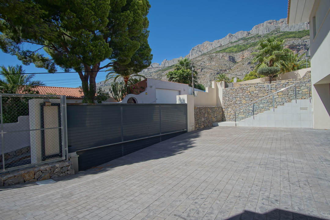 Main entrance featuring a spacious stamped concrete driveway, dark sliding gate, and natural stone walls set against the Sierra de Altea mountains.