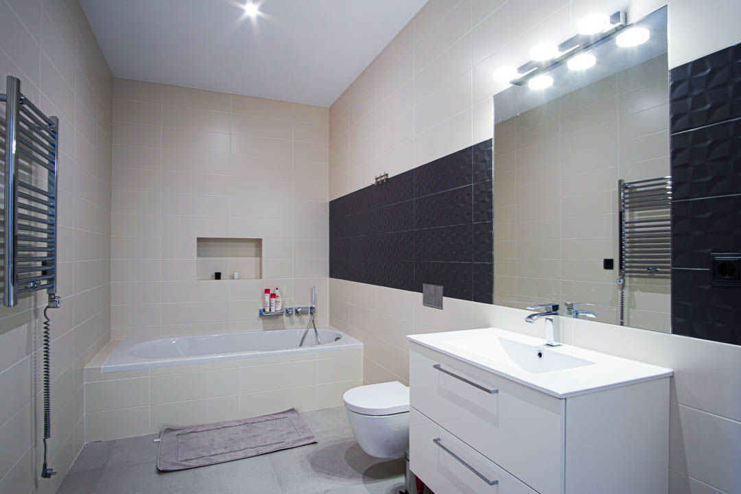 Modern bathroom featuring a built-in bathtub, white vanity with dual drawers, wall-mounted toilet, and beige wall tiles with a textured dark grey accent strip.