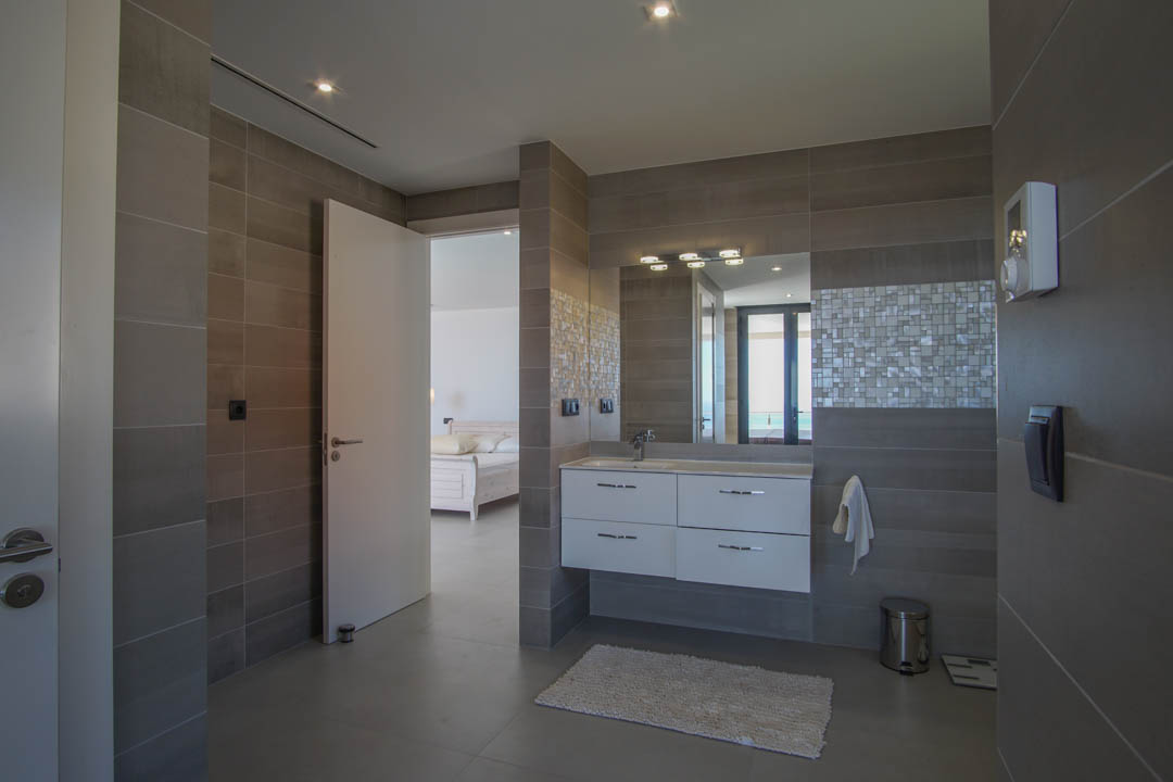 Modern bathroom featuring grey tiling, a white floating vanity, and mosaic accents. The open doorway reveals the master suite and distant Mediterranean views.