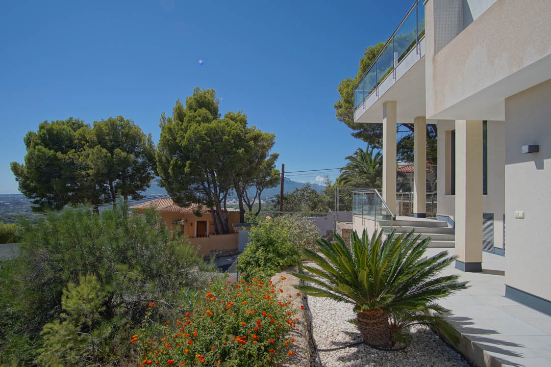 Modern exterior facade featuring white columns, glass railings, and a landscaped garden with gravel, a cycad palm, and Mediterranean flora.